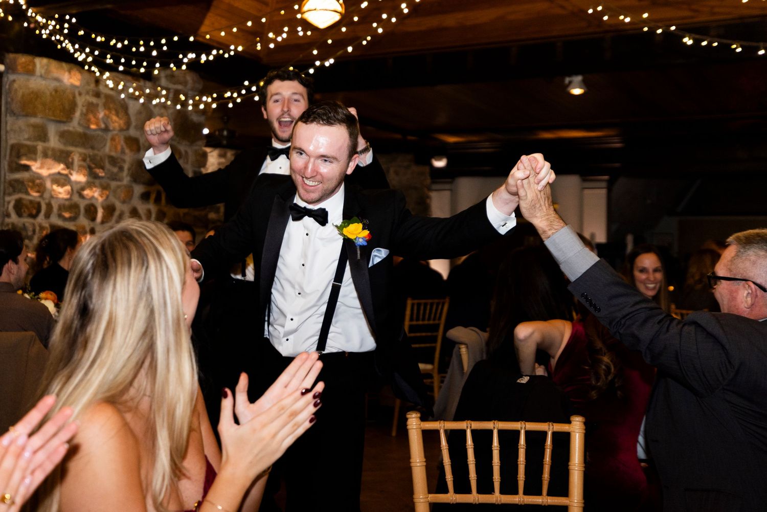 Groomsmen in tuxedo dancing and celebrating with guests at wedding reception under string lights.