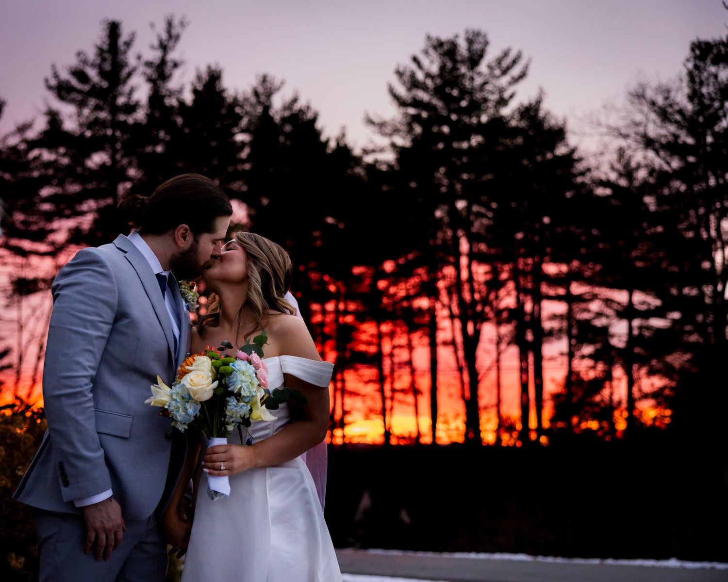 A couple shares a passionate kiss silhouetted against a breathtaking fiery sunset with pine trees behind them.
