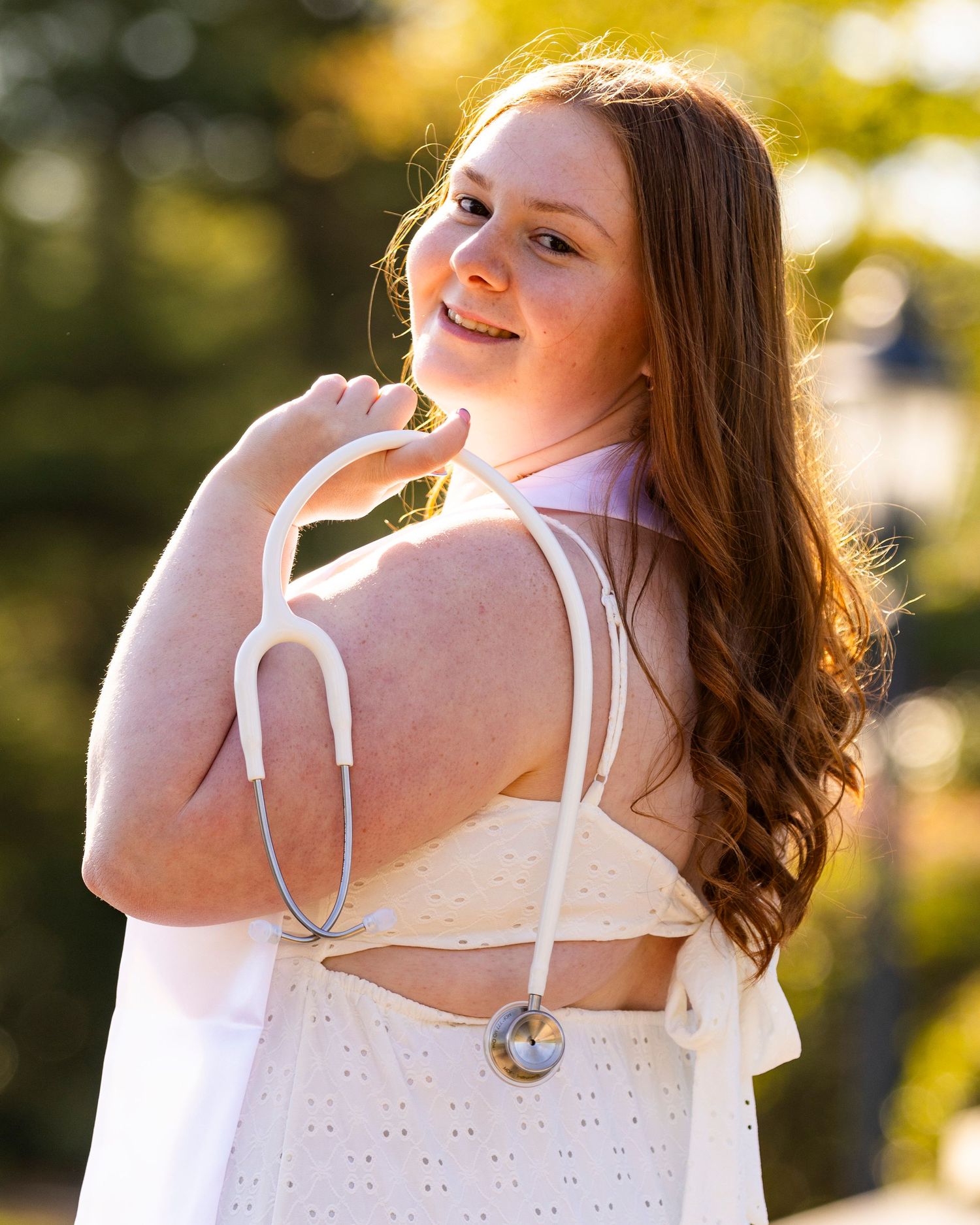 A young woman with long red hair poses outdoors in a white dress with a stethoscope draped over her shoulder.