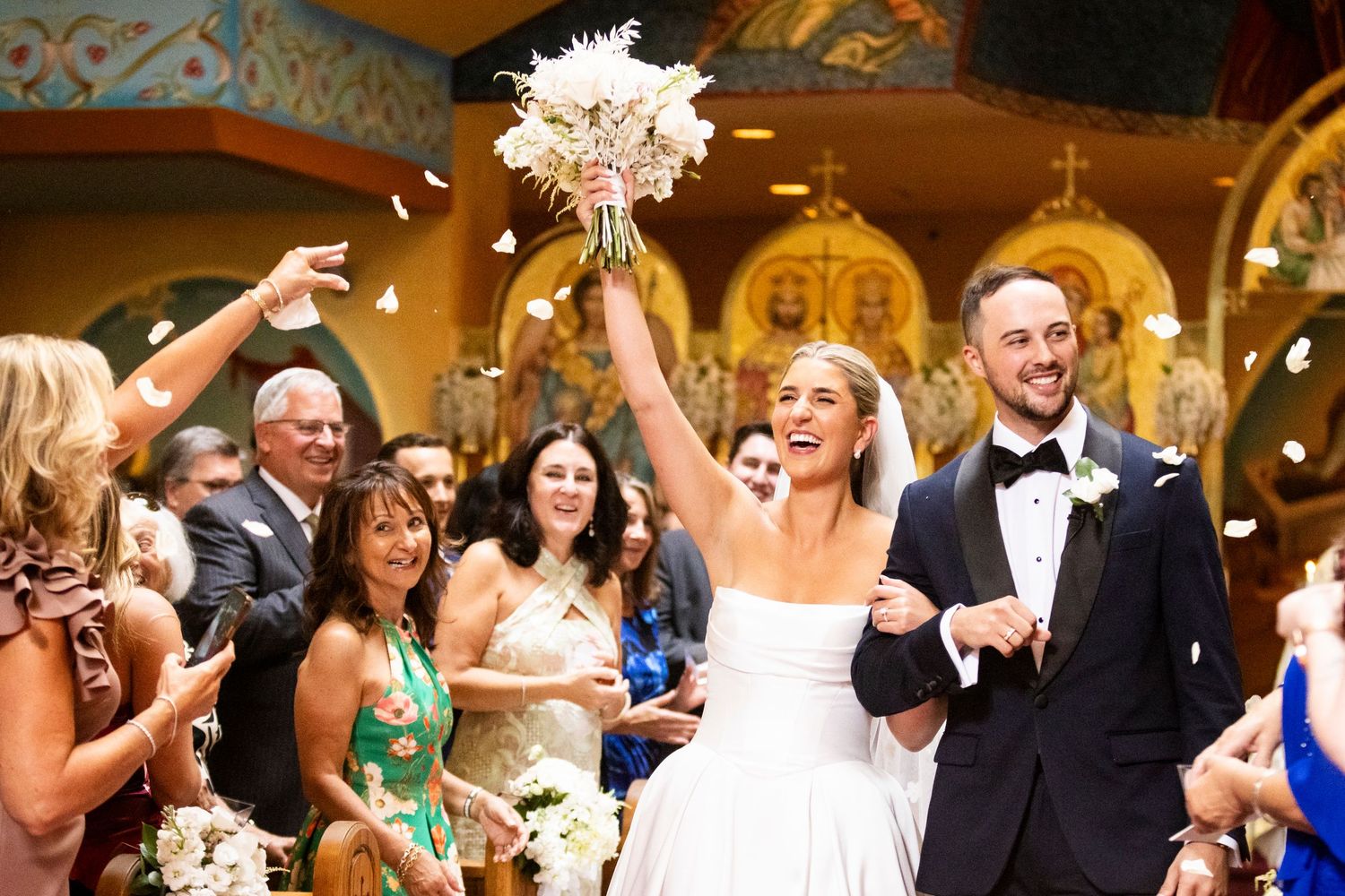 Joyful bride raises bouquet triumphantly as couple exits church ceremony while guests celebrate with confetti.