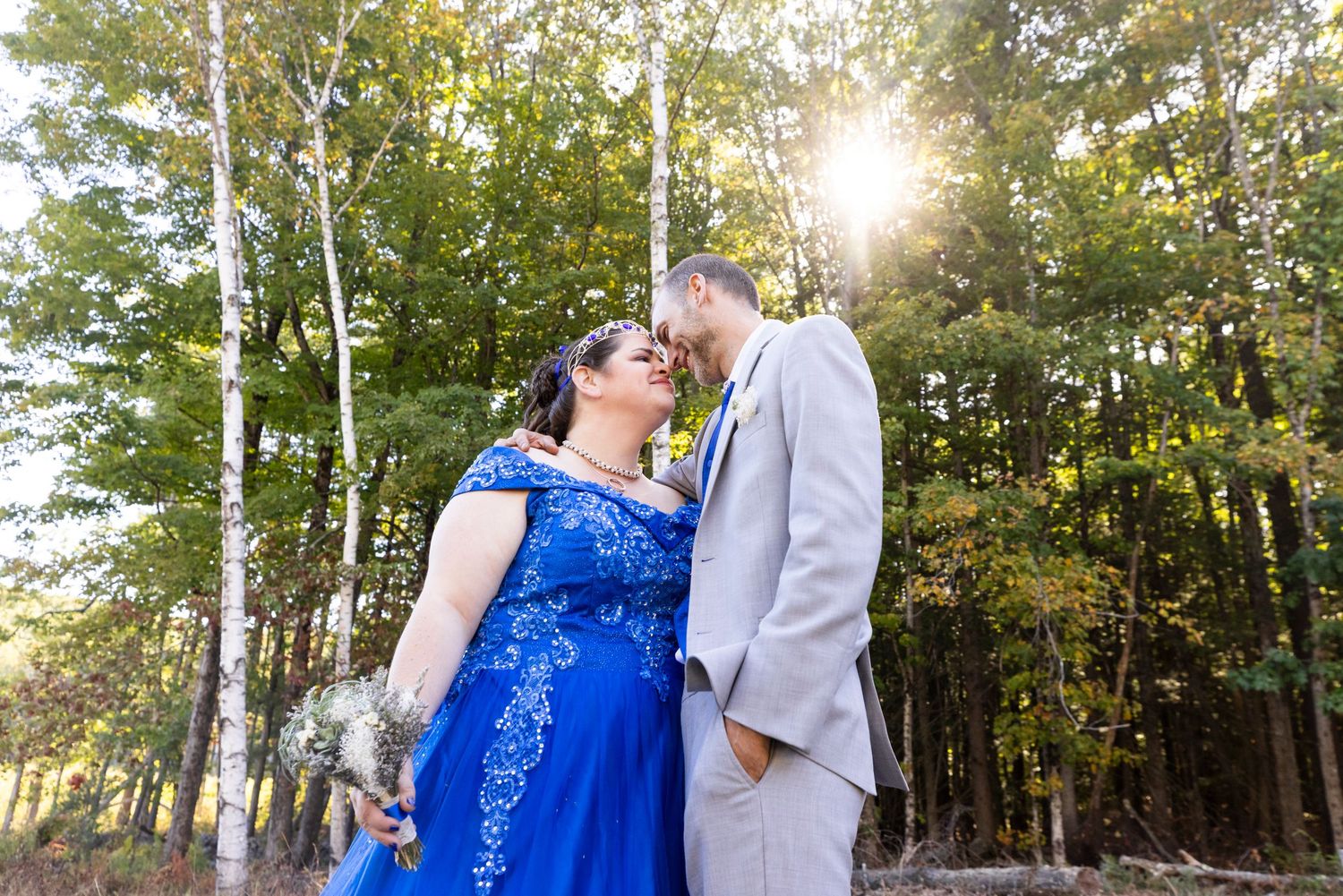 Couple sharing romantic kiss in sunlit forest clearing, bride in royal blue wedding dress.