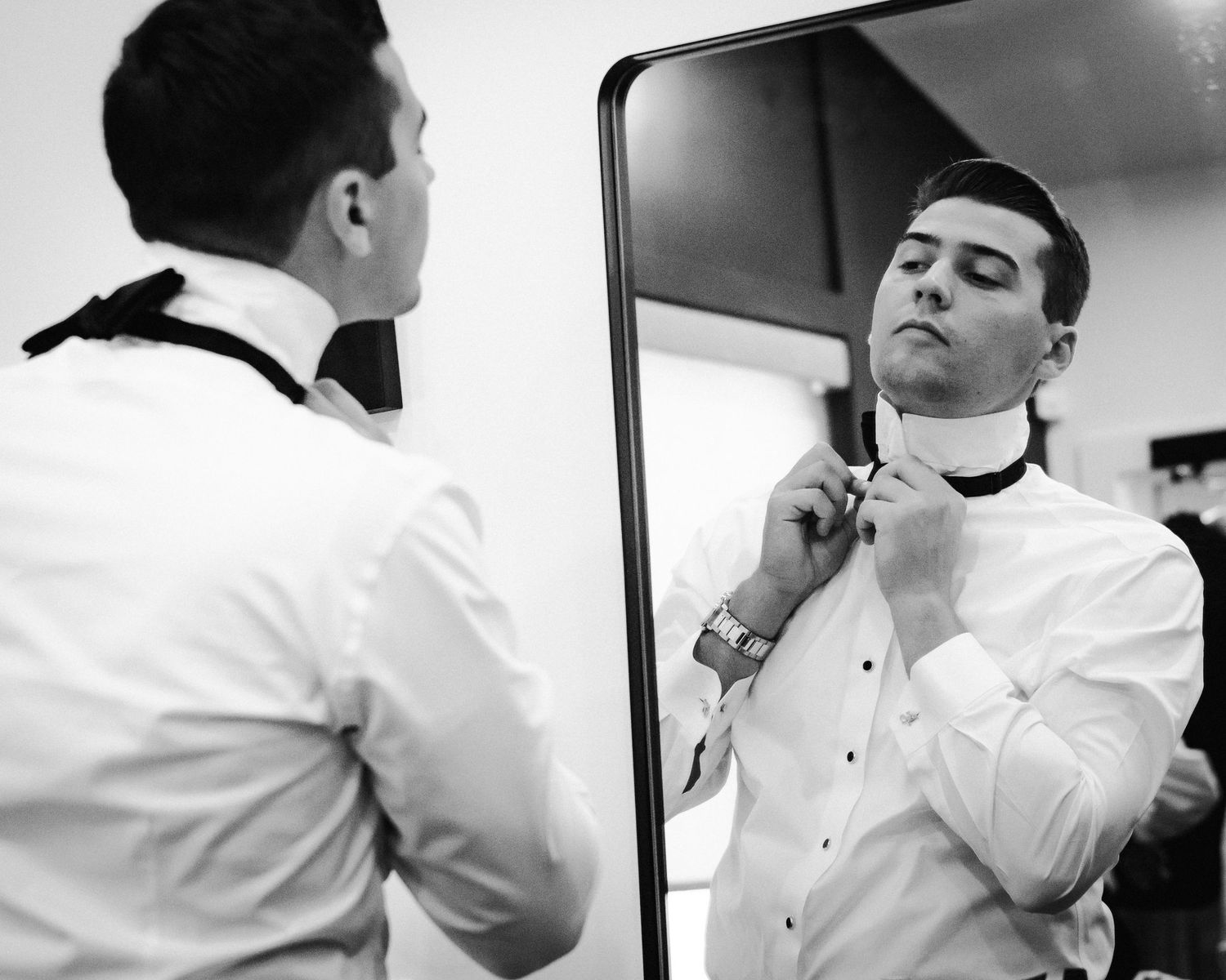 Groom adjusting his bow tie while looking in a mirror, black and white wedding preparation photo.