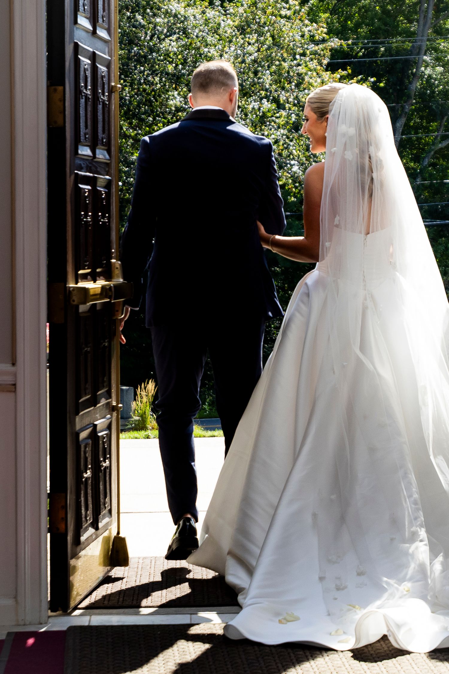 Bride in flowing white gown and veil following groom through ornate church entrance doors.