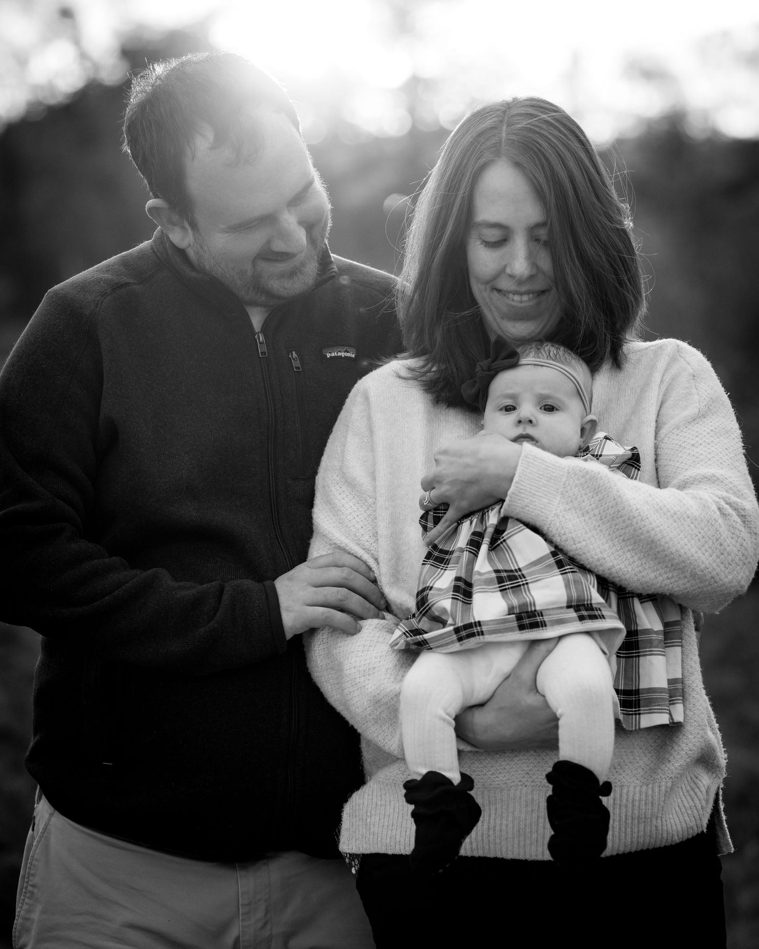 Black and white portrait of smiling parents holding their newborn baby outdoors at sunset.
