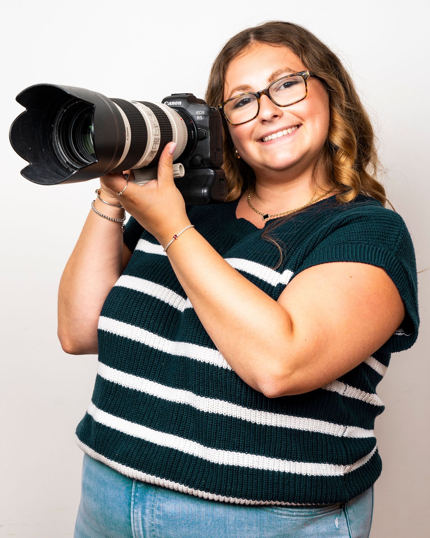 A smiling female photographer poses holding a Canon DSLR camera with telephoto lens indoors.