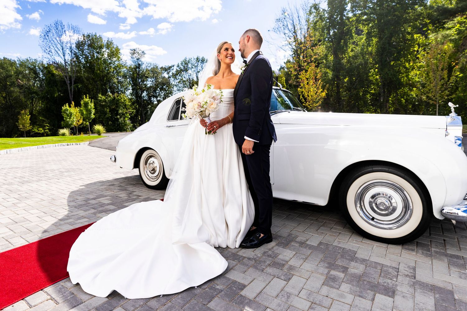 An elegant bride and groom pose beside a classic white vintage Rolls Royce on a red carpet outdoors.