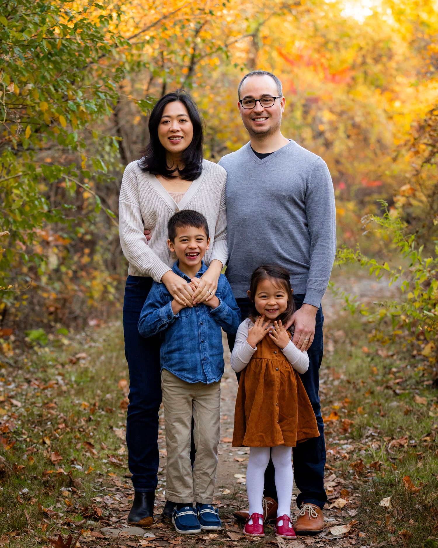 A smiling family of four poses together on a scenic autumn path with vibrant fall foliage in the background.