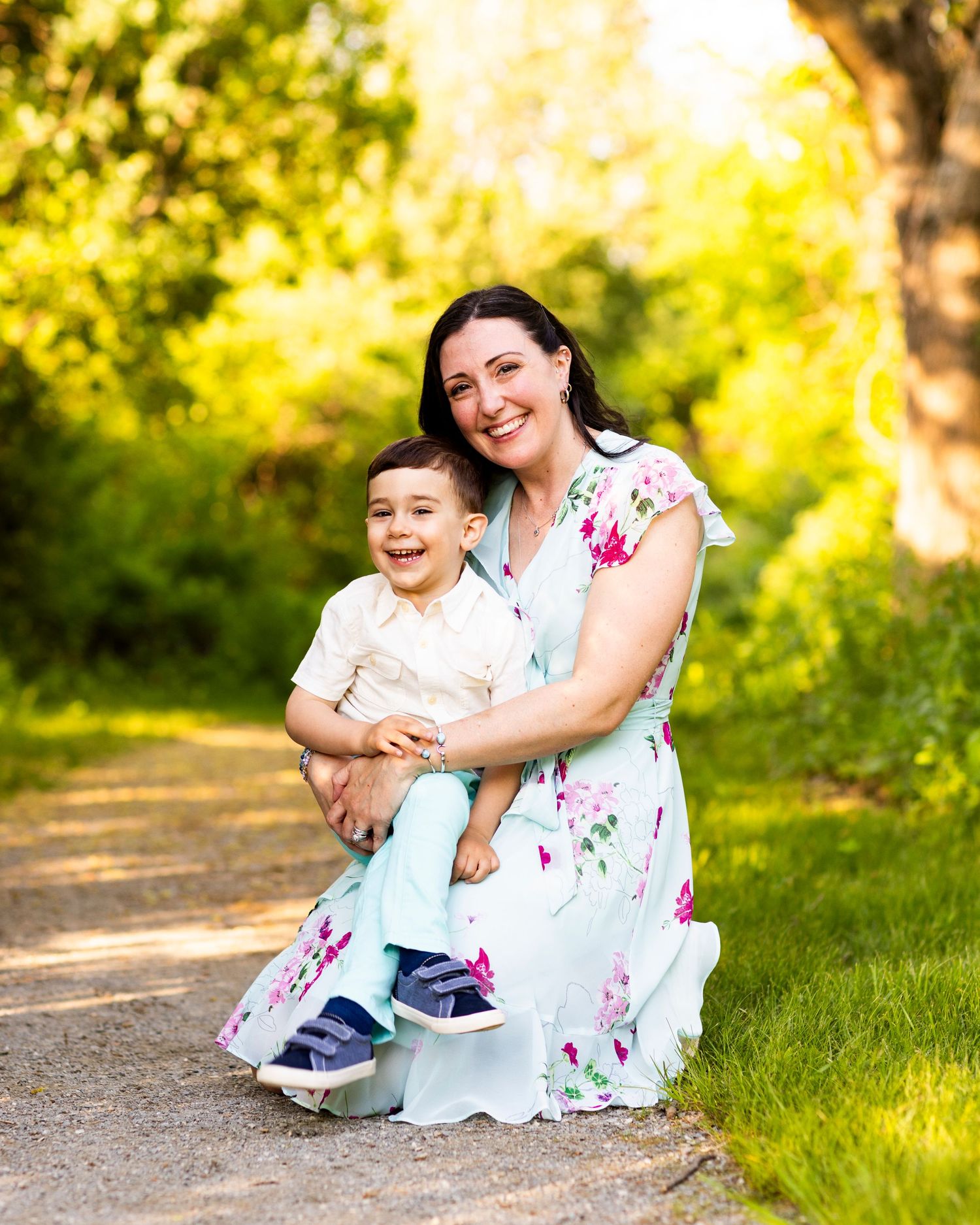 Smiling mother in floral dress sitting on garden path holding her laughing toddler son in golden light.
