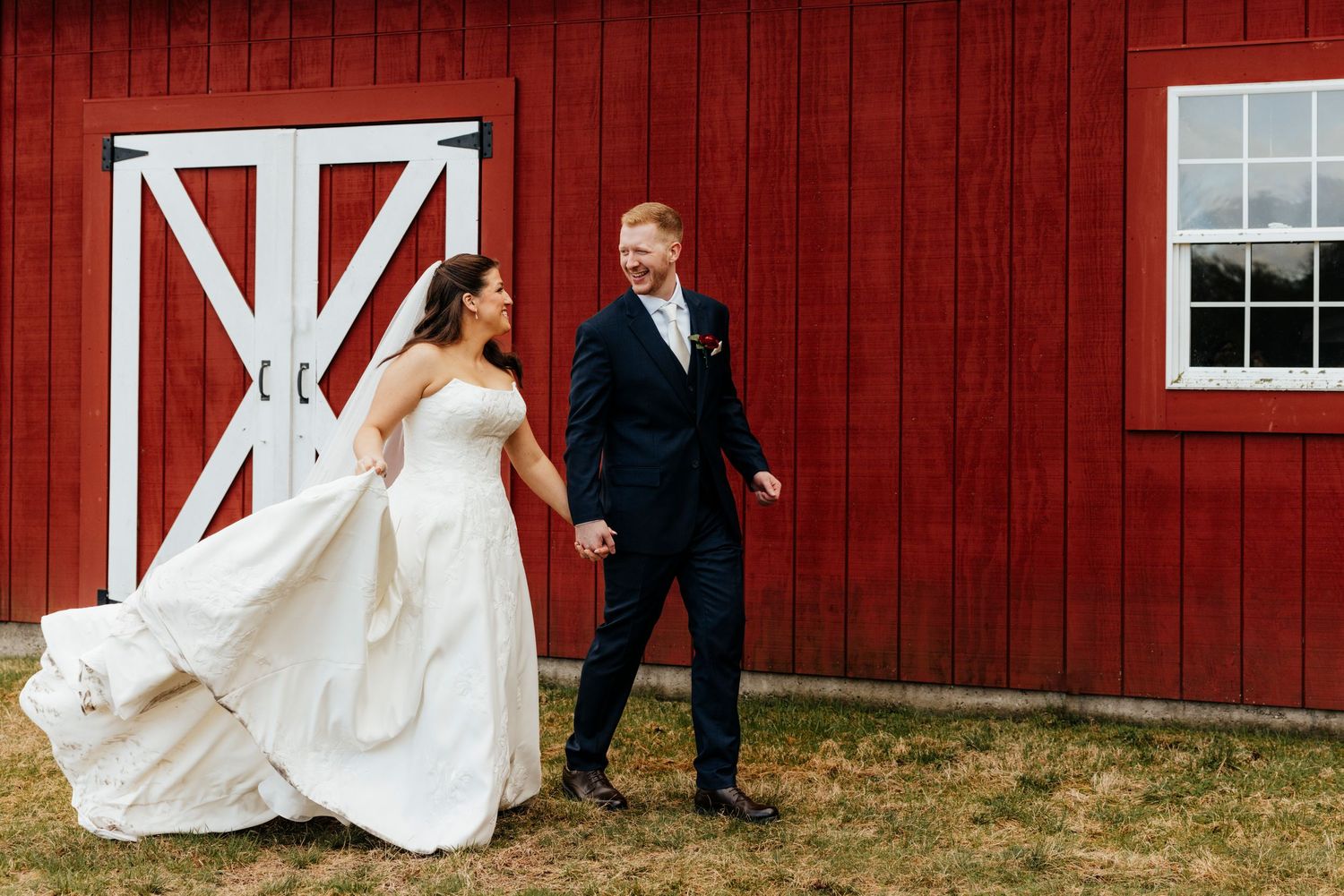 Bride and groom walk hand-in-hand laughing beside a red barn on their wedding day at Publick House in