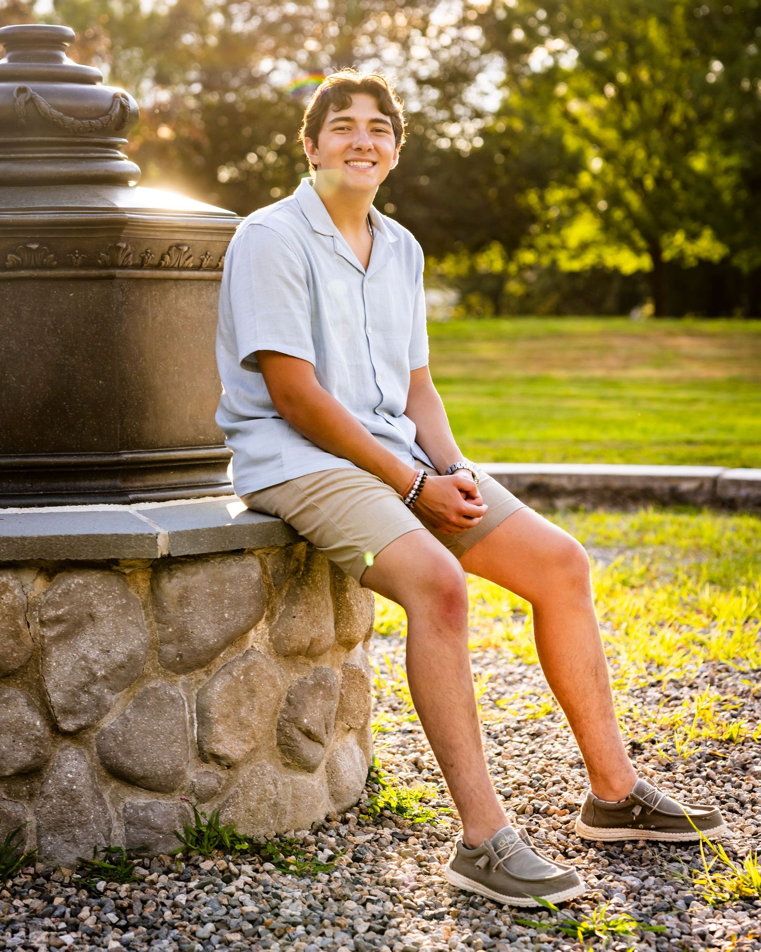Young man in light blue shirt and khaki shorts sitting on stone wall in sunny park for senior photos.