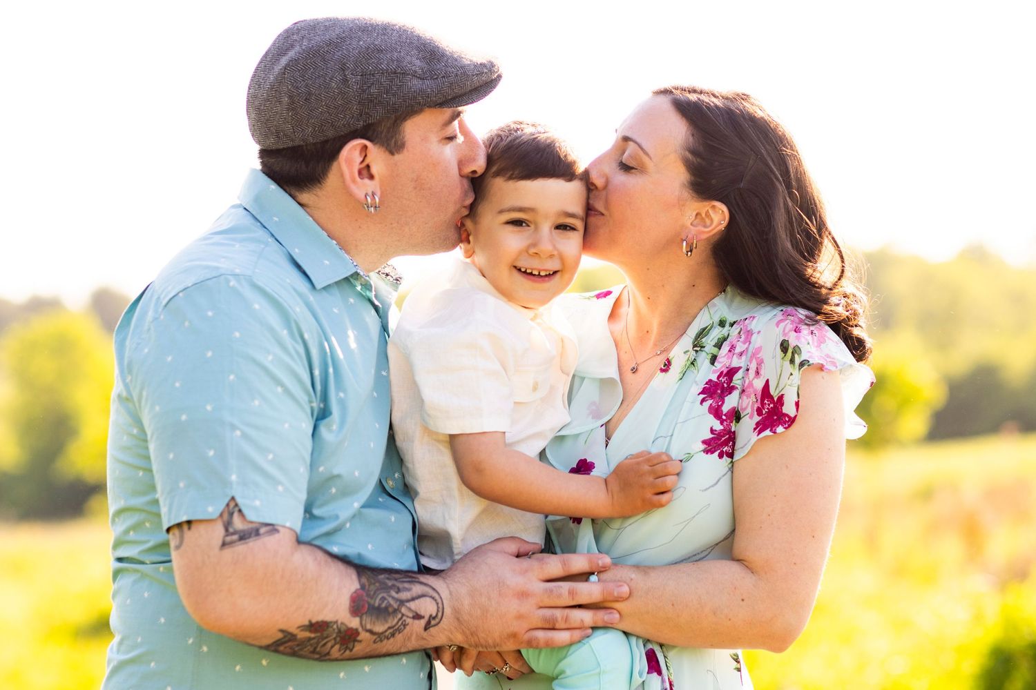 Happy parents kissing their laughing baby boy outdoors in a sunny golden field setting.
