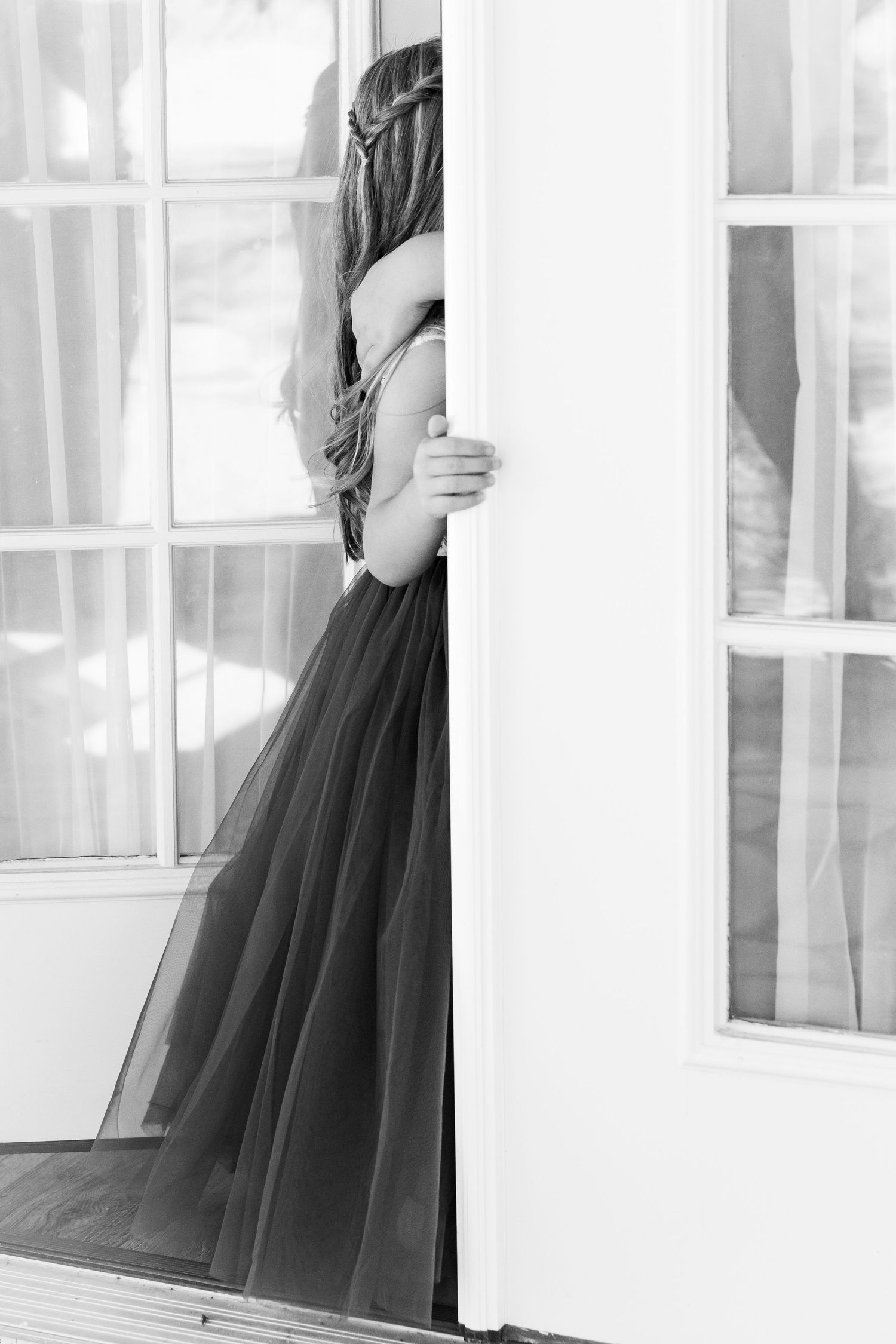Bride in elegant gown peeks around white French door in black and white photo.
