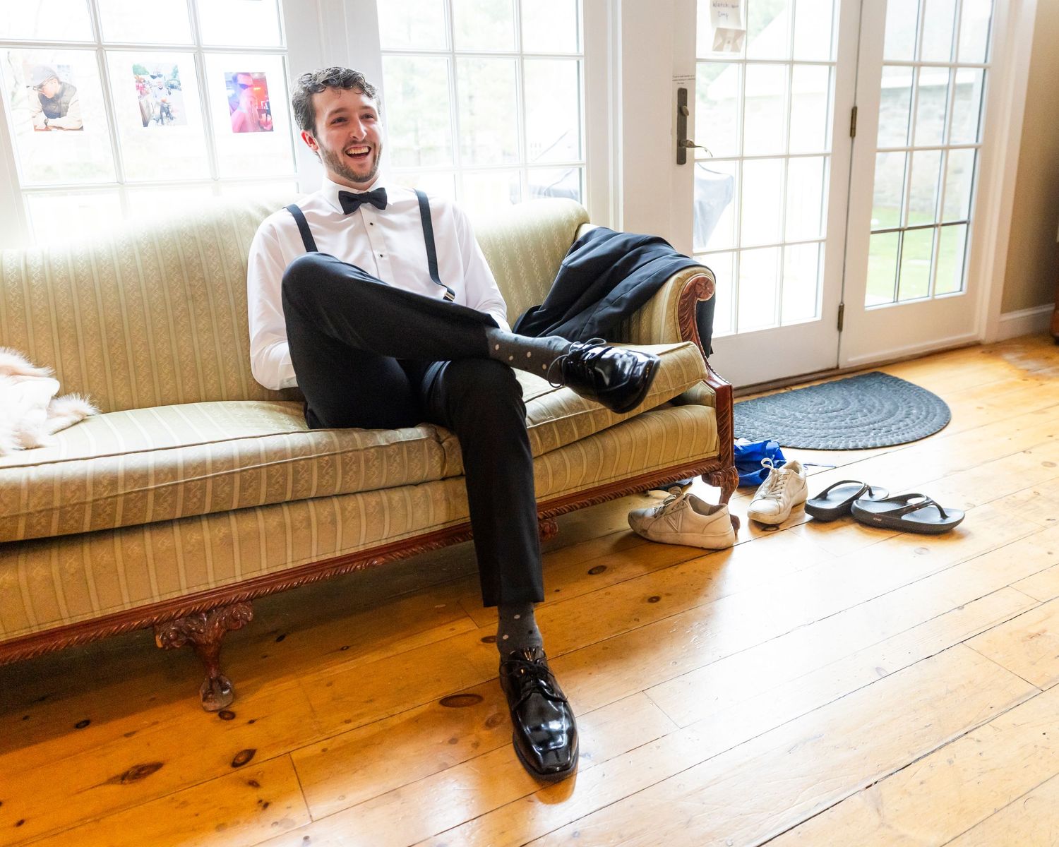 Relaxed groom in suspenders and bow tie smiling on a couch while getting ready for his wedding.