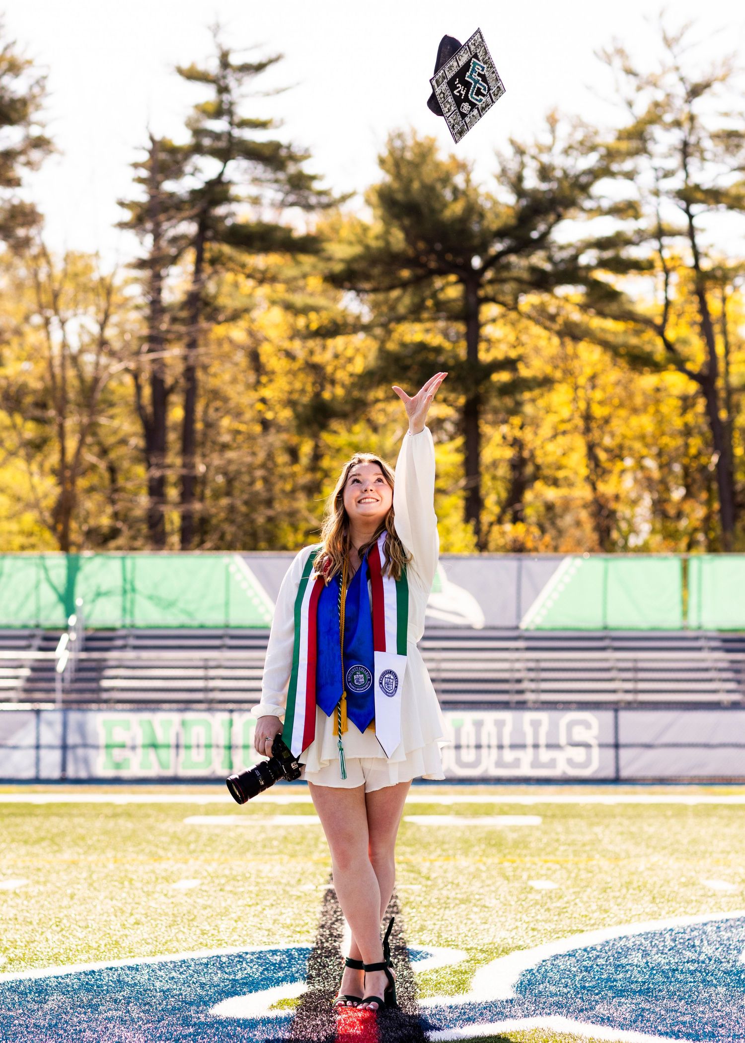 A graduate tosses her cap in the air on a football field wearing a white dress and colorful graduation stoles.