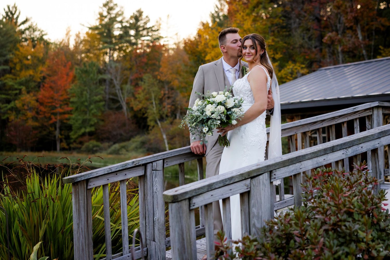 Bride and groom pose on rustic wooden bridge with vibrant fall foliage backdrop.