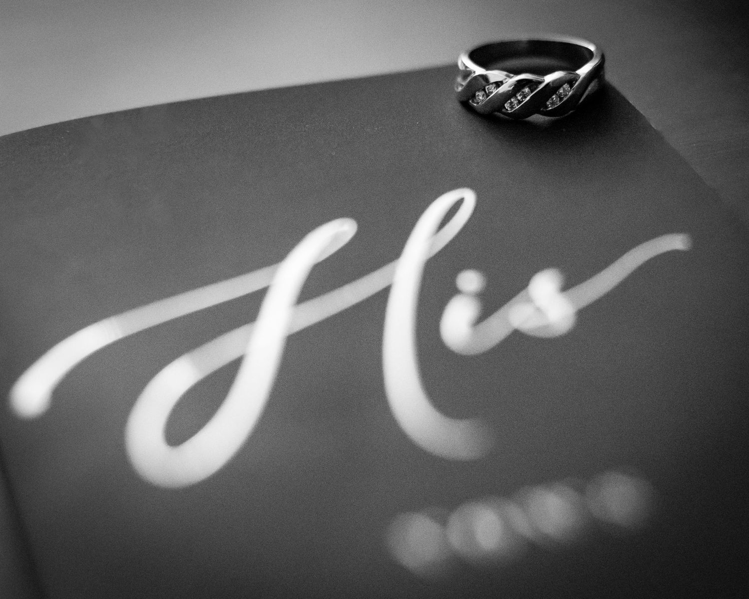Close-up black and white photo of a diamond ring resting on a book labeled His.