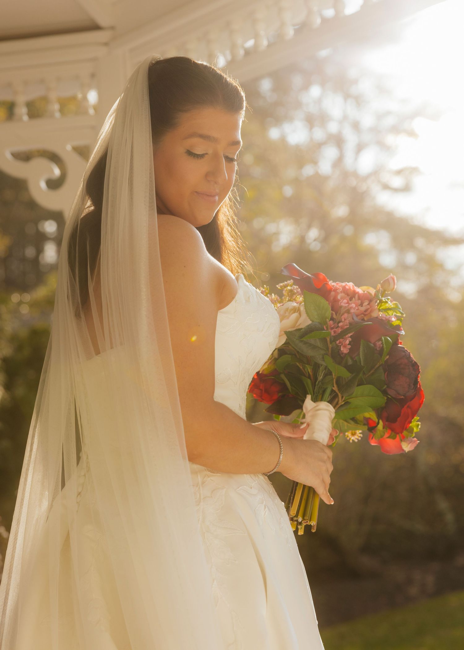 Bride in white gown and veil holds colorful bouquet of red flowers, bathed in golden sunlight outdoors at The Publick House Historic Inn in