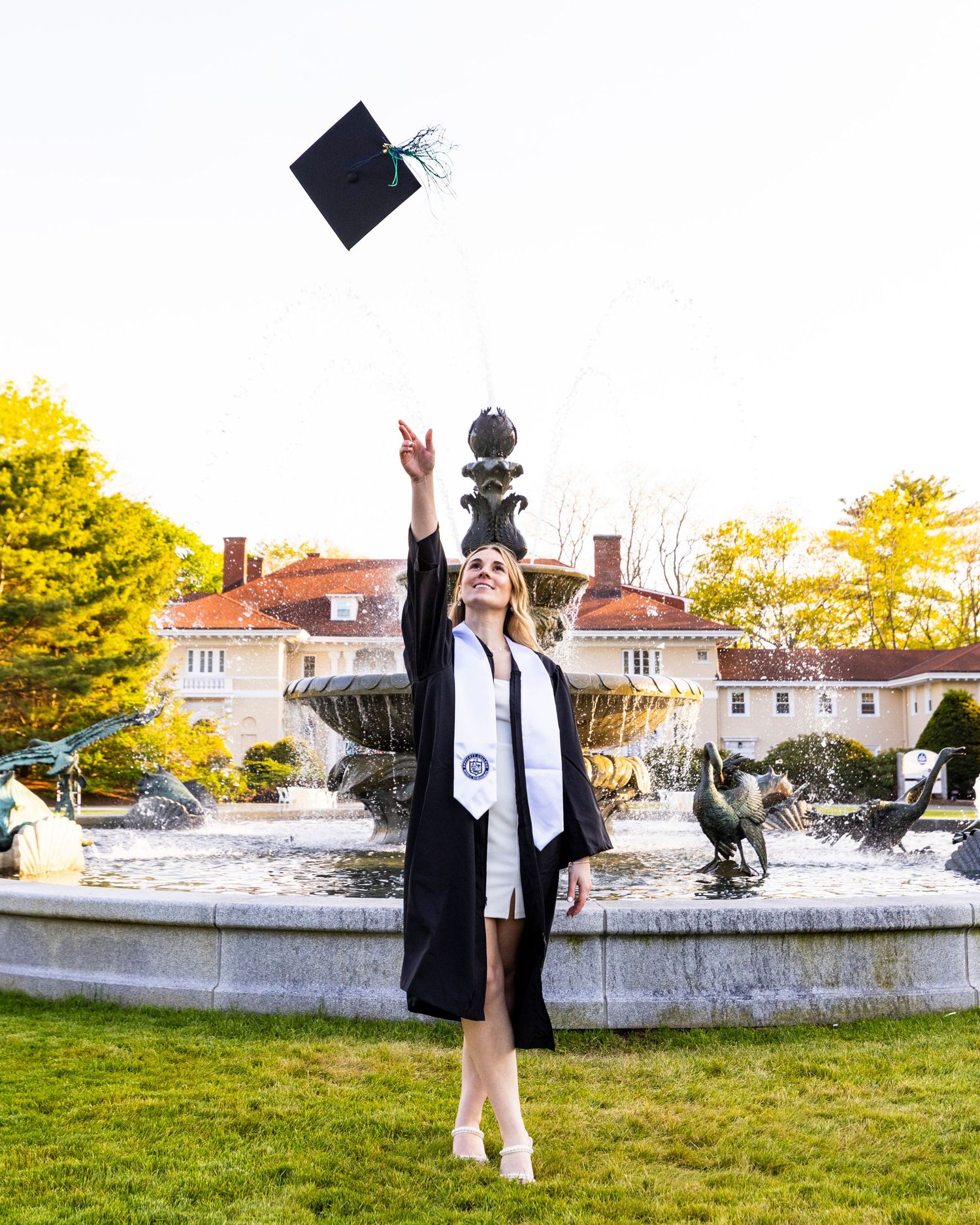A graduate in cap and gown tosses her mortarboard into the air by a fountain during an outdoor graduation photo session.