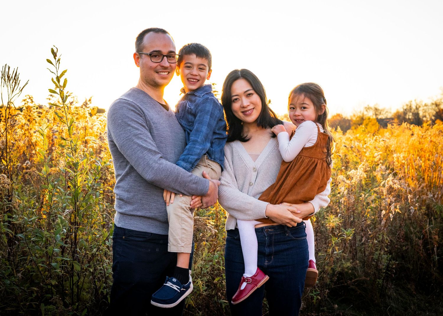 A happy family of four poses together in a sunlit autumn field during an outdoor photo session.