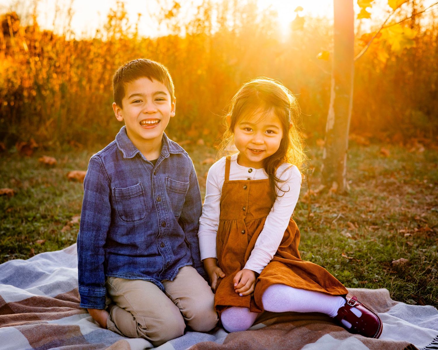 A smiling brother and sister sit together on a blanket outdoors in a beautiful golden autumn setting.