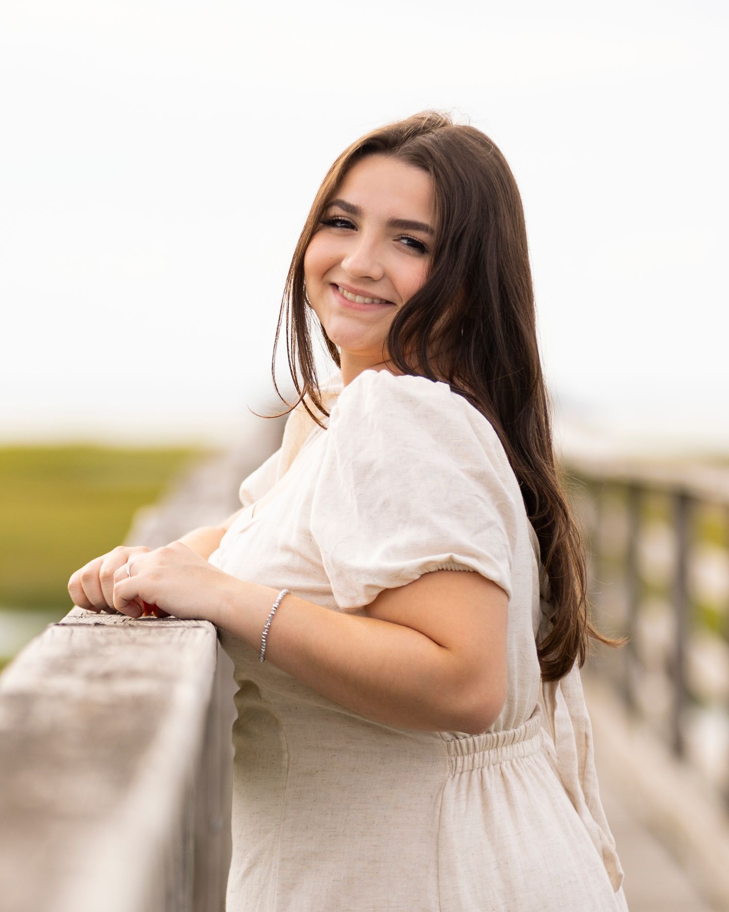Smiling brunette teen girl in white dress leaning on wooden railing for senior portrait outdoors.