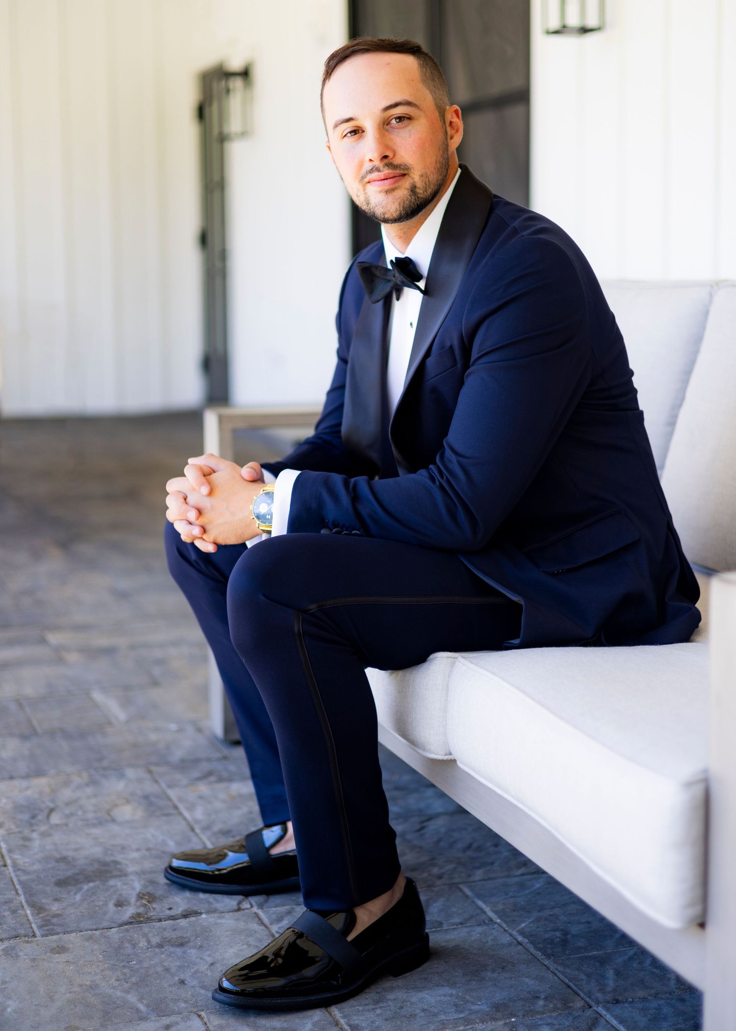 Groom in navy tuxedo with bow tie and black loafers seated on white outdoor bench.