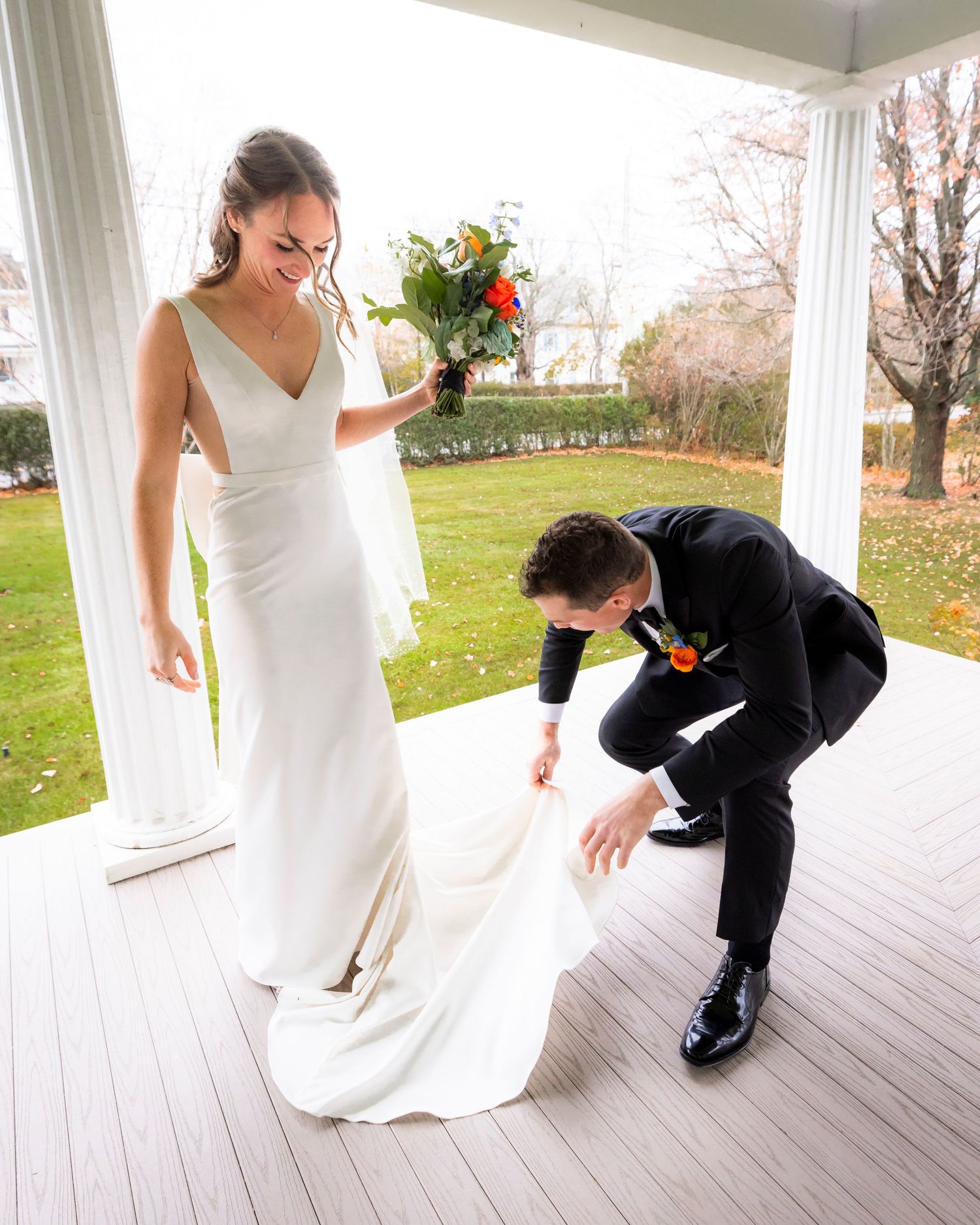 Groom adjusting bride's wedding dress train on a porch while she holds a colorful bouquet.
