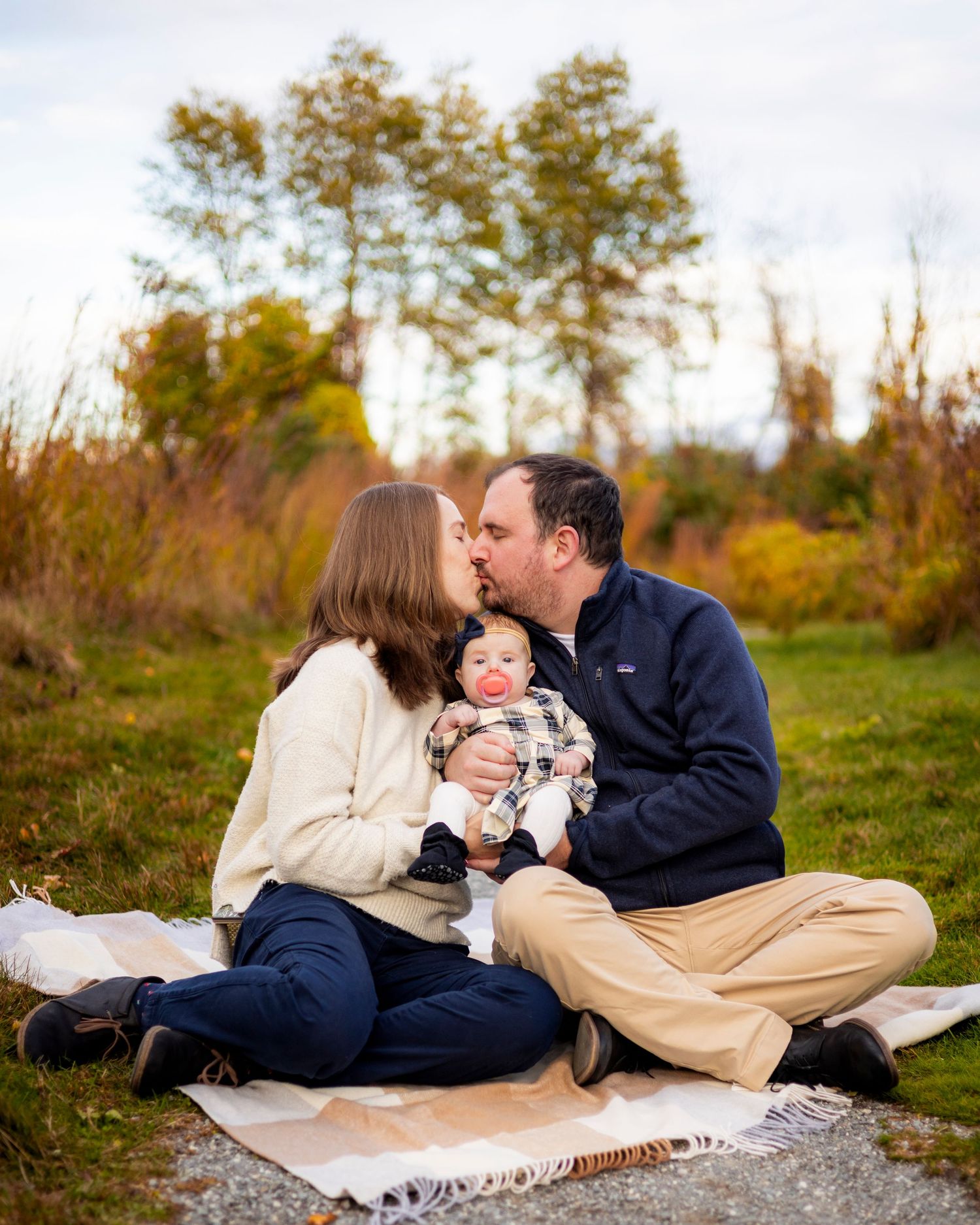 New parents share a kiss while holding their baby on a blanket during an autumn outdoor session.