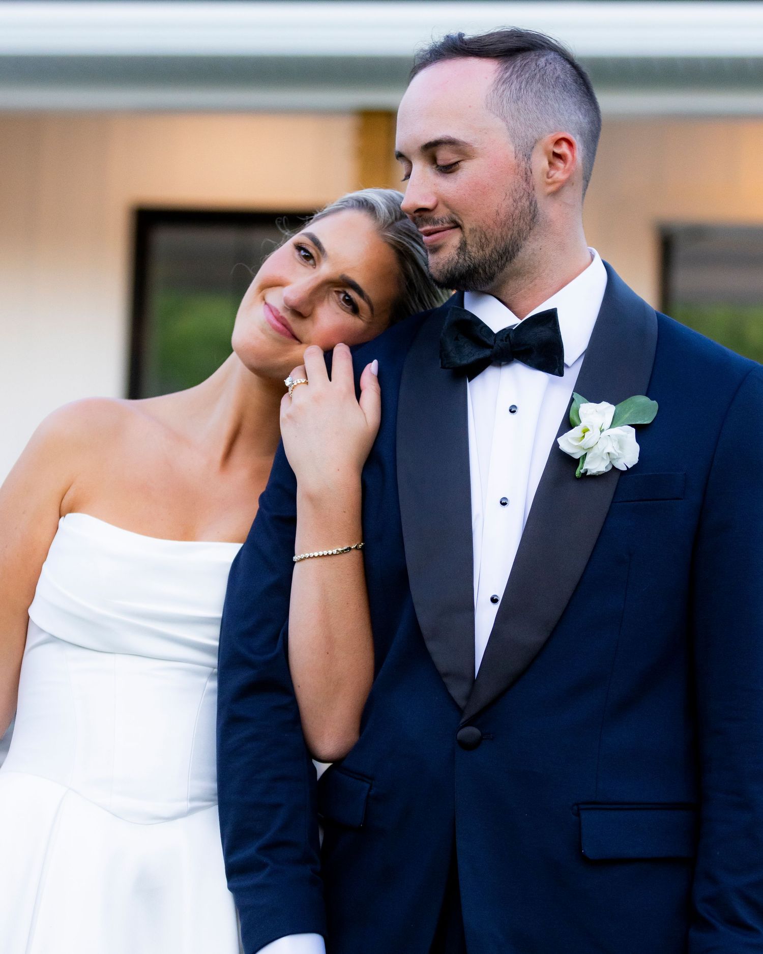 Bride leans head on groom's shoulder as he closes his eyes during romantic outdoor wedding portrait.
