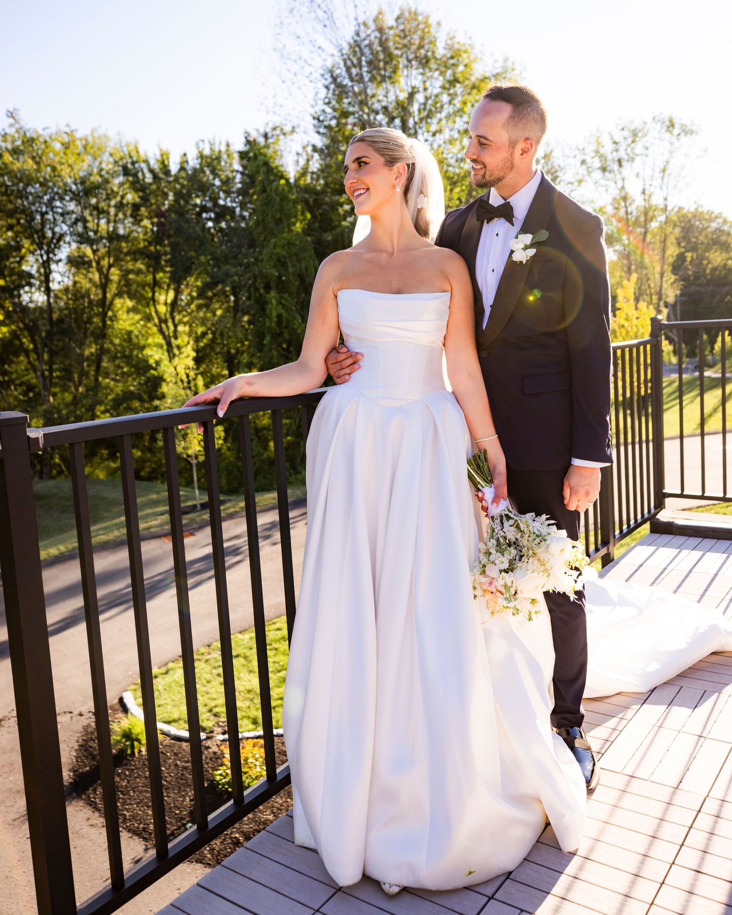 Elegant bride in strapless gown holds bouquet beside tuxedo-clad groom on sunny balcony.