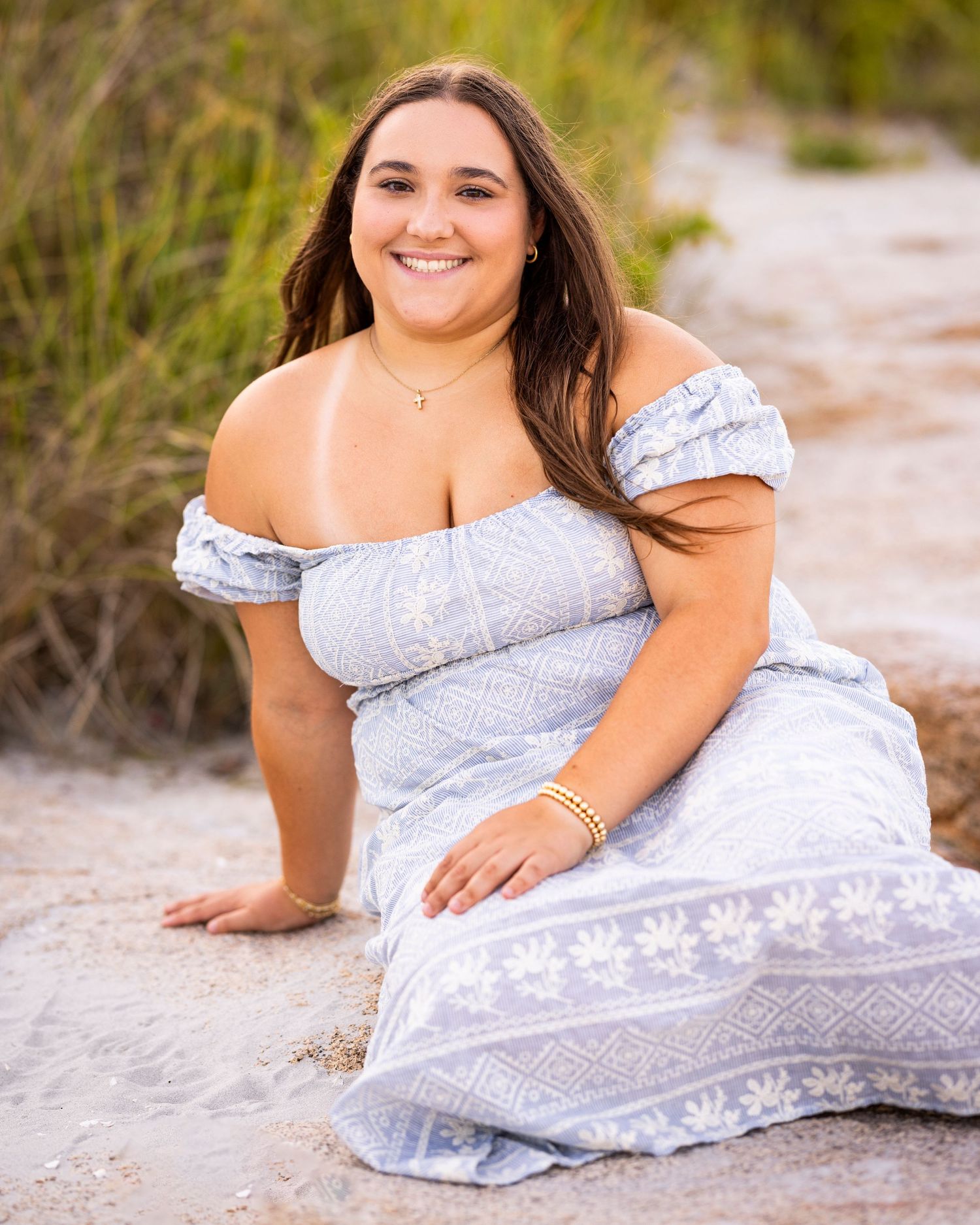 Smiling young woman in blue off-shoulder floral dress poses on sandy beach at sunset.
