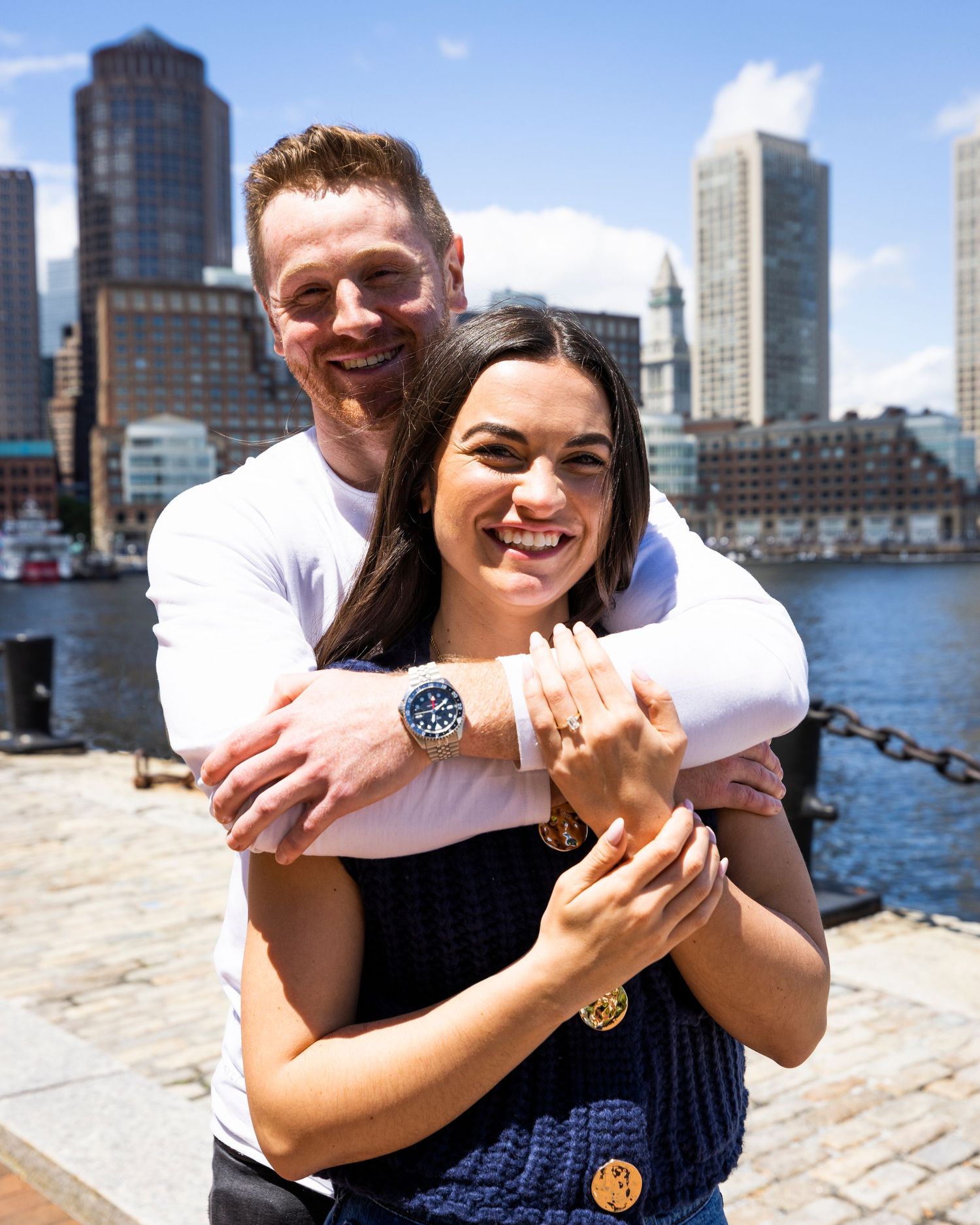 Happy newly engaged couple embracing on Boston waterfront, woman showing engagement ring with city skyline behind.
