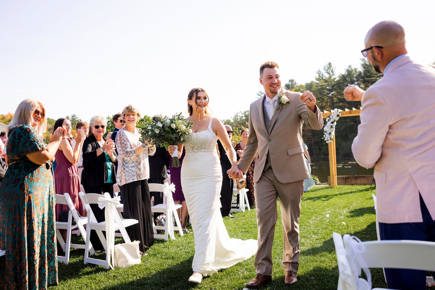 Bride and groom walk joyfully down the aisle at outdoor wedding ceremony with guests applauding.