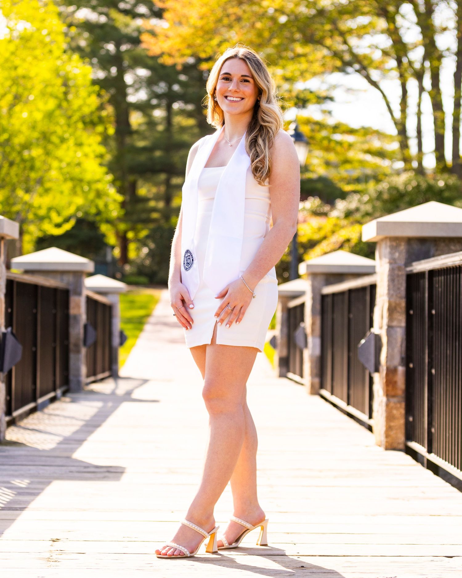 Smiling blonde woman in white dress standing on bridge surrounded by fall foliage for graduation photos.
