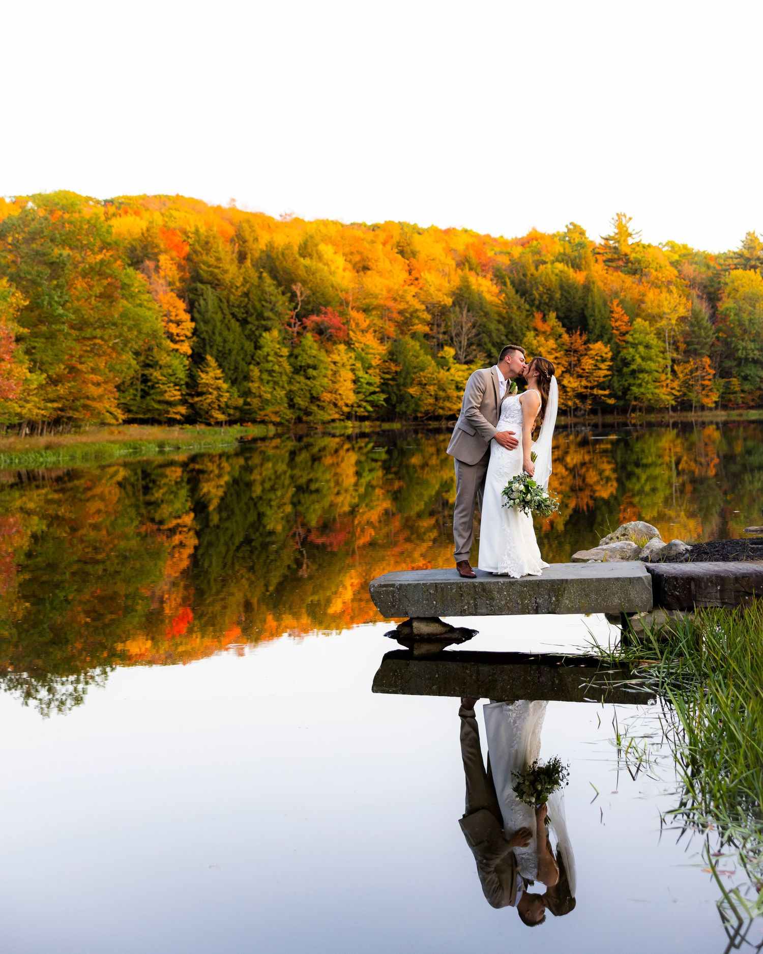 Couple kissing on a dock by a reflective lake surrounded by vibrant fall foliage during an autumn wedding.