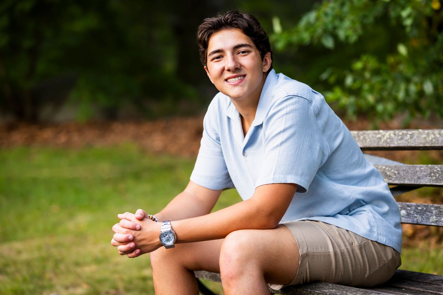 Smiling young man in light blue shirt sitting on wooden bench outdoors for senior portrait session.