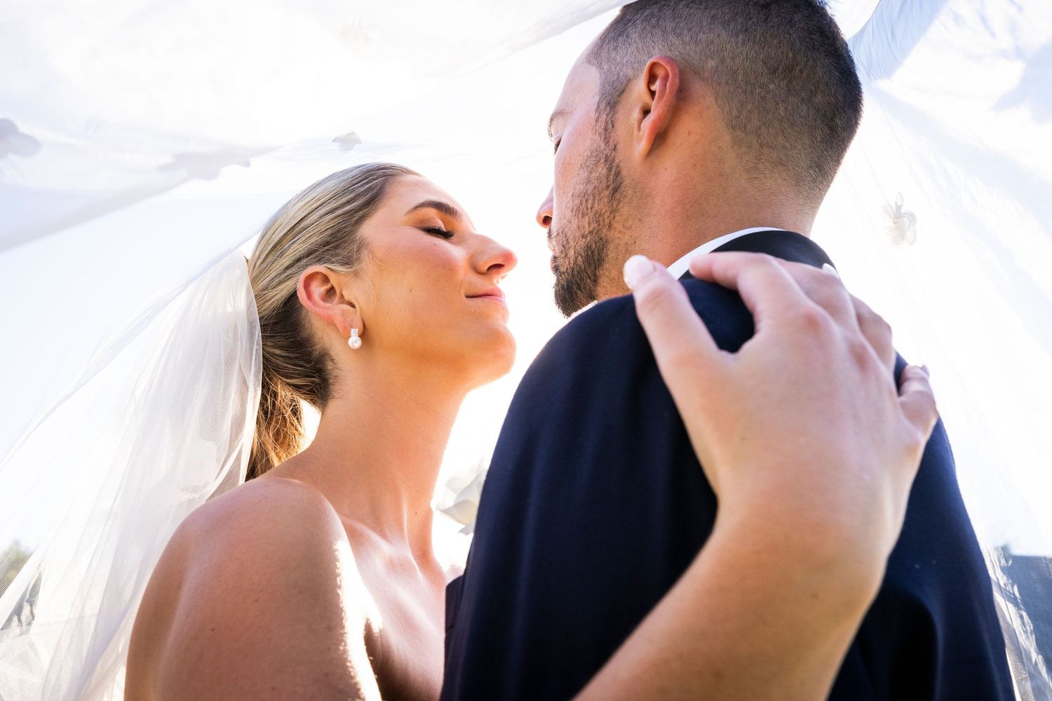 Romantic bride and groom sharing an intimate moment under a flowing white veil in soft natural light.