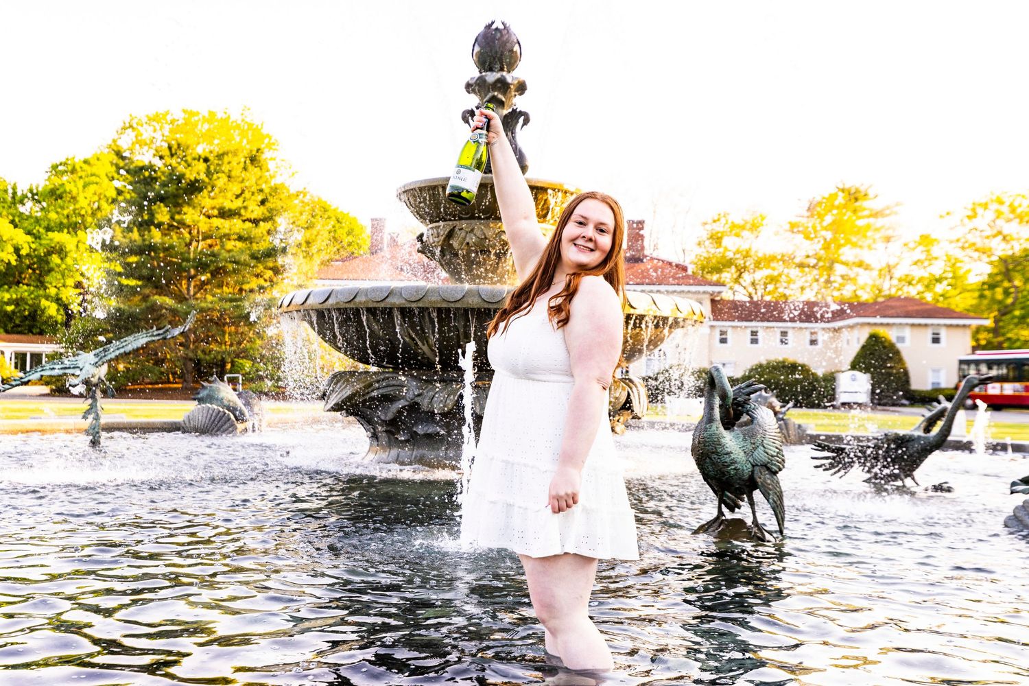A young woman in a white dress celebrates in a fountain, holding a champagne bottle aloft during a graduation photo shoot.
