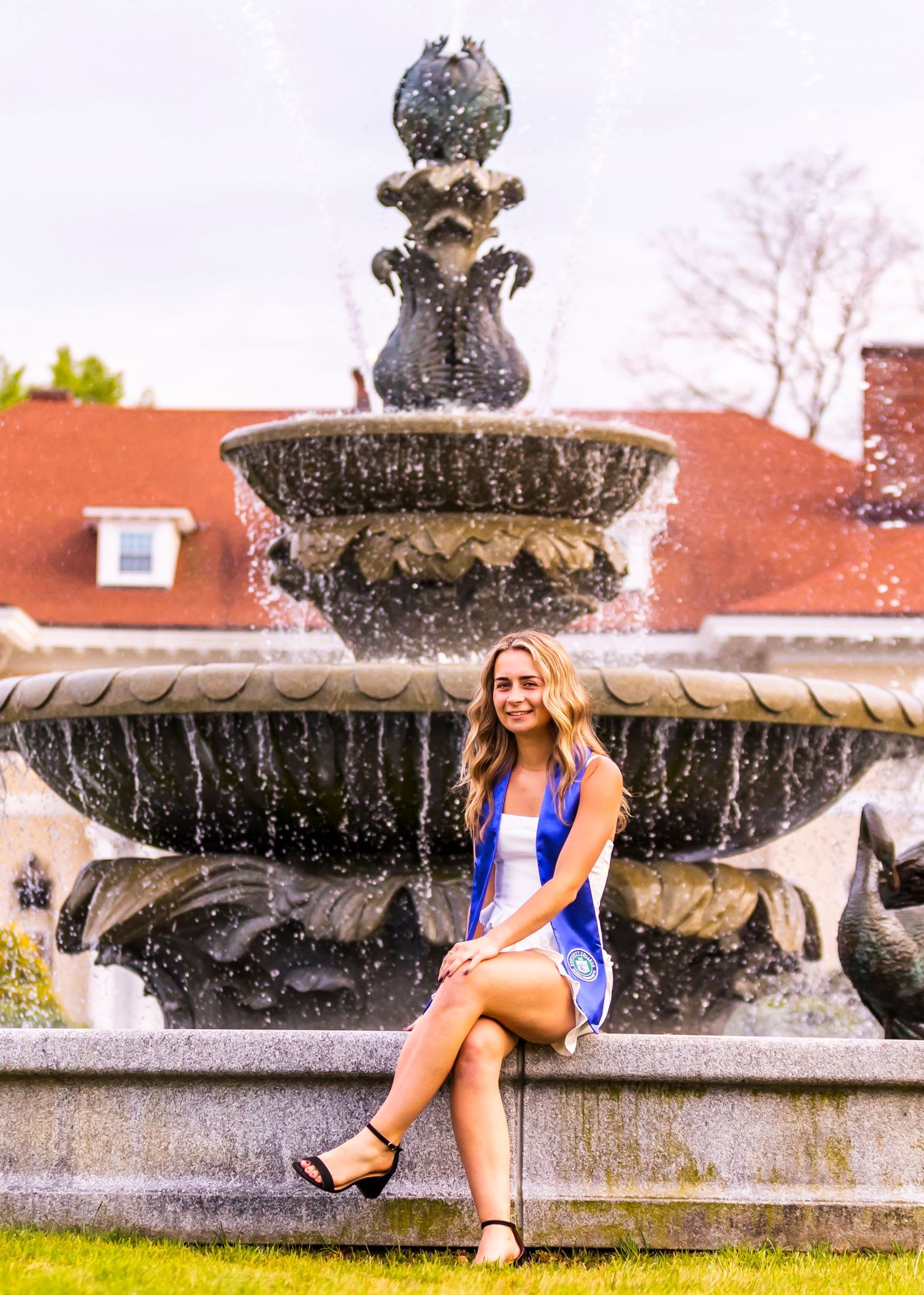 Female graduate in blue dress sitting by ornate fountain on college campus for graduation photos.