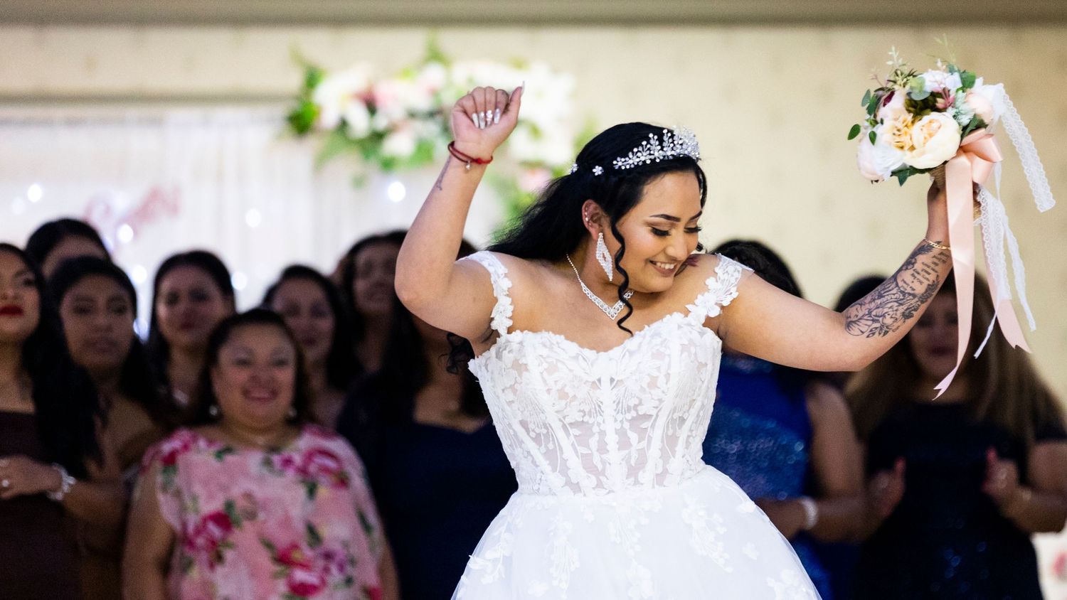 Bride in white lace gown celebrating with raised arms while tossing bouquet at wedding reception.
