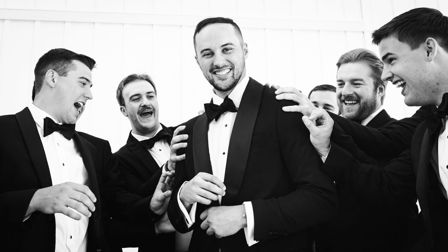 Black and white image of groom and groomsmen in tuxedos sharing laughs before wedding ceremony.