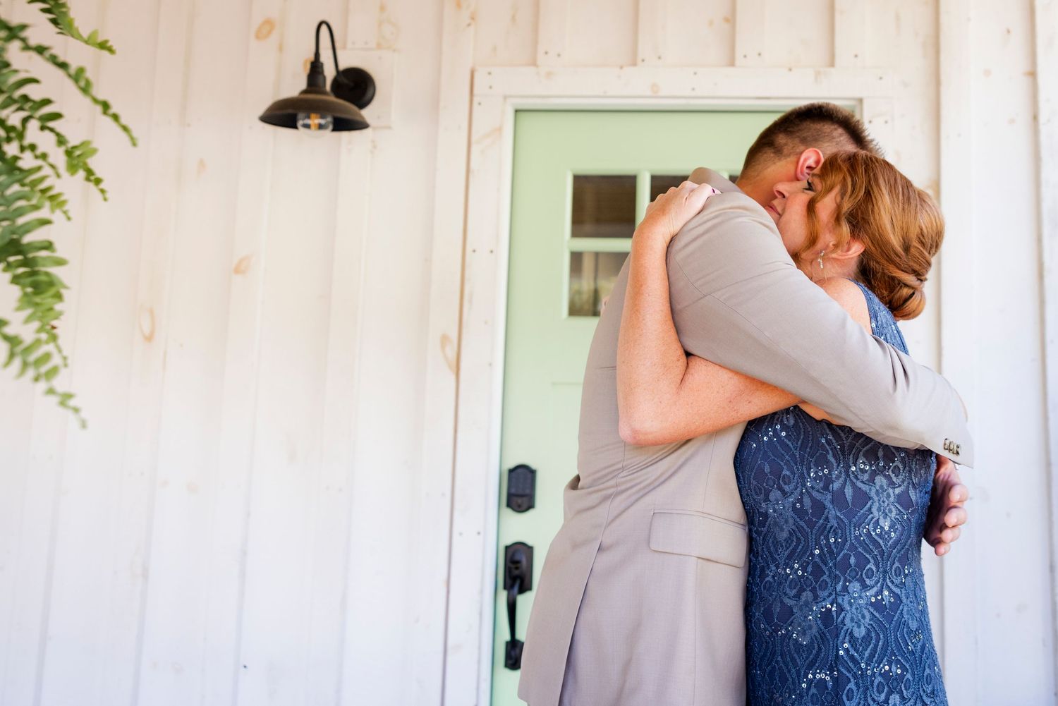 Groom and woman in blue sequin dress sharing a warm embrace outside white barn door.