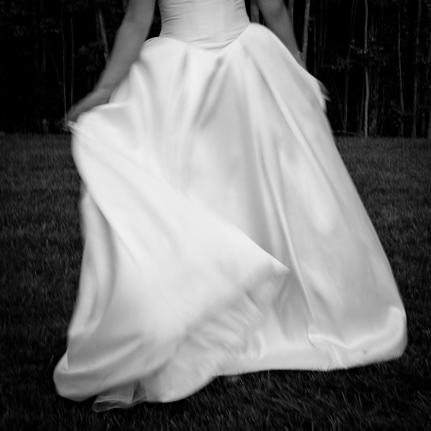 Bride twirls flowing white ball gown skirt on grass in artistic black and white photo.