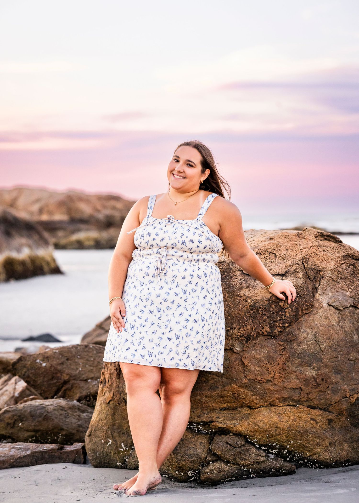 Young woman in a white floral dress poses against coastal rocks during a stunning pink sunset senior portrait session.