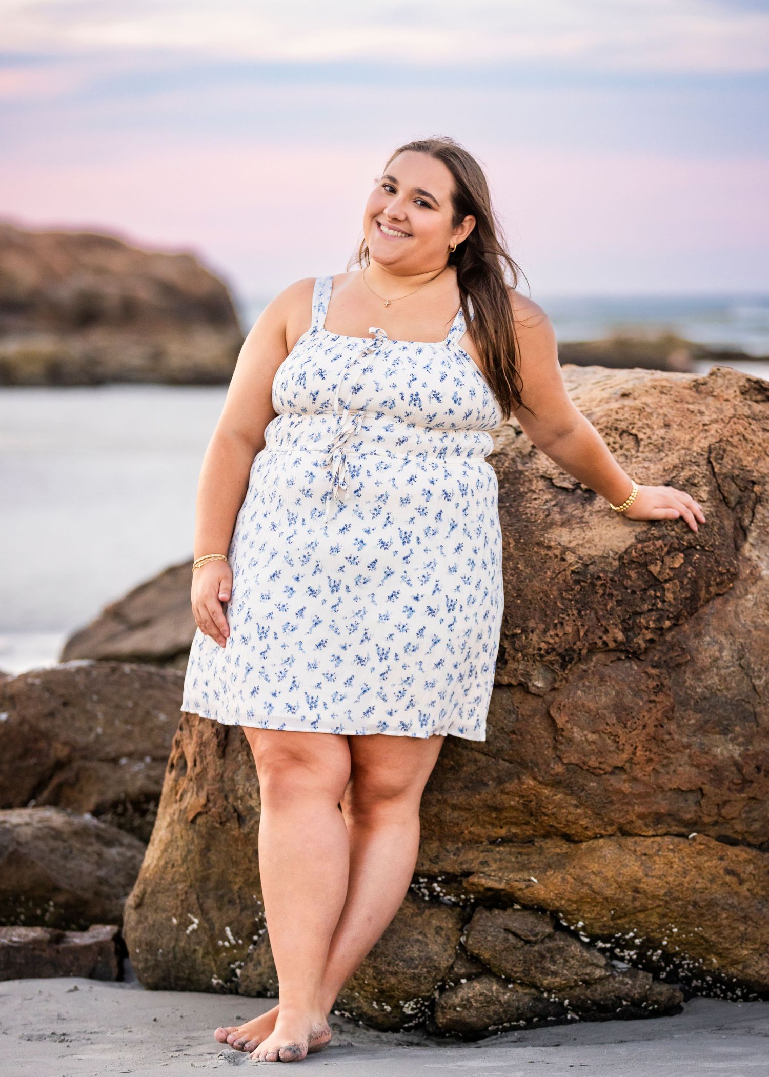 Smiling young woman in a white floral sundress posing against coastal rocks with a pastel sunset sky.