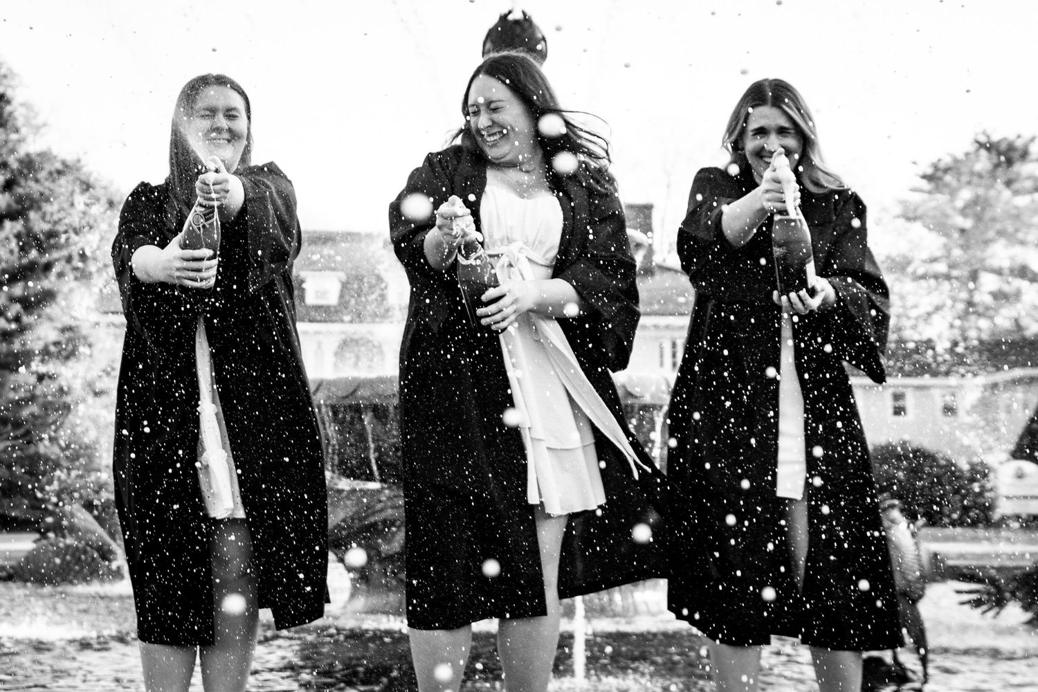 Three women in black coats celebrating with champagne bottles in black and white photo outdoors.