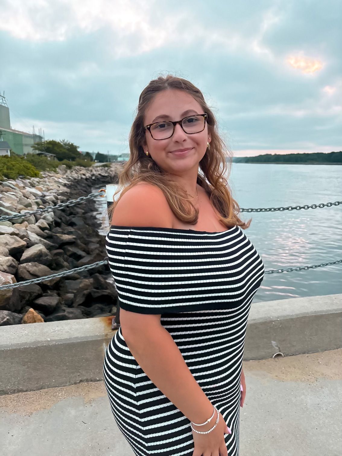 A young woman in a black and white striped off-shoulder dress poses by a waterfront rocky shoreline.