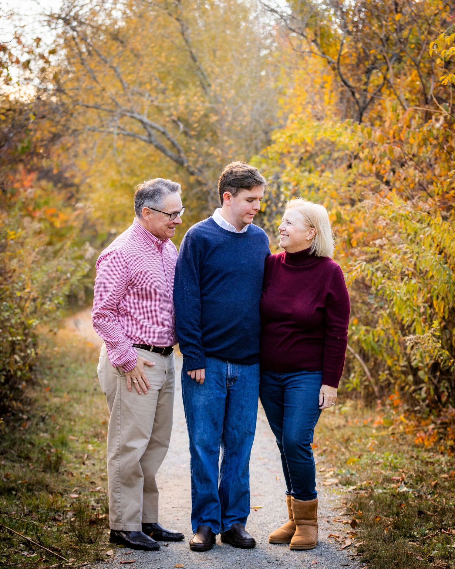 Three people smile together on an autumn trail surrounded by colorful fall foliage during a family portrait session.
