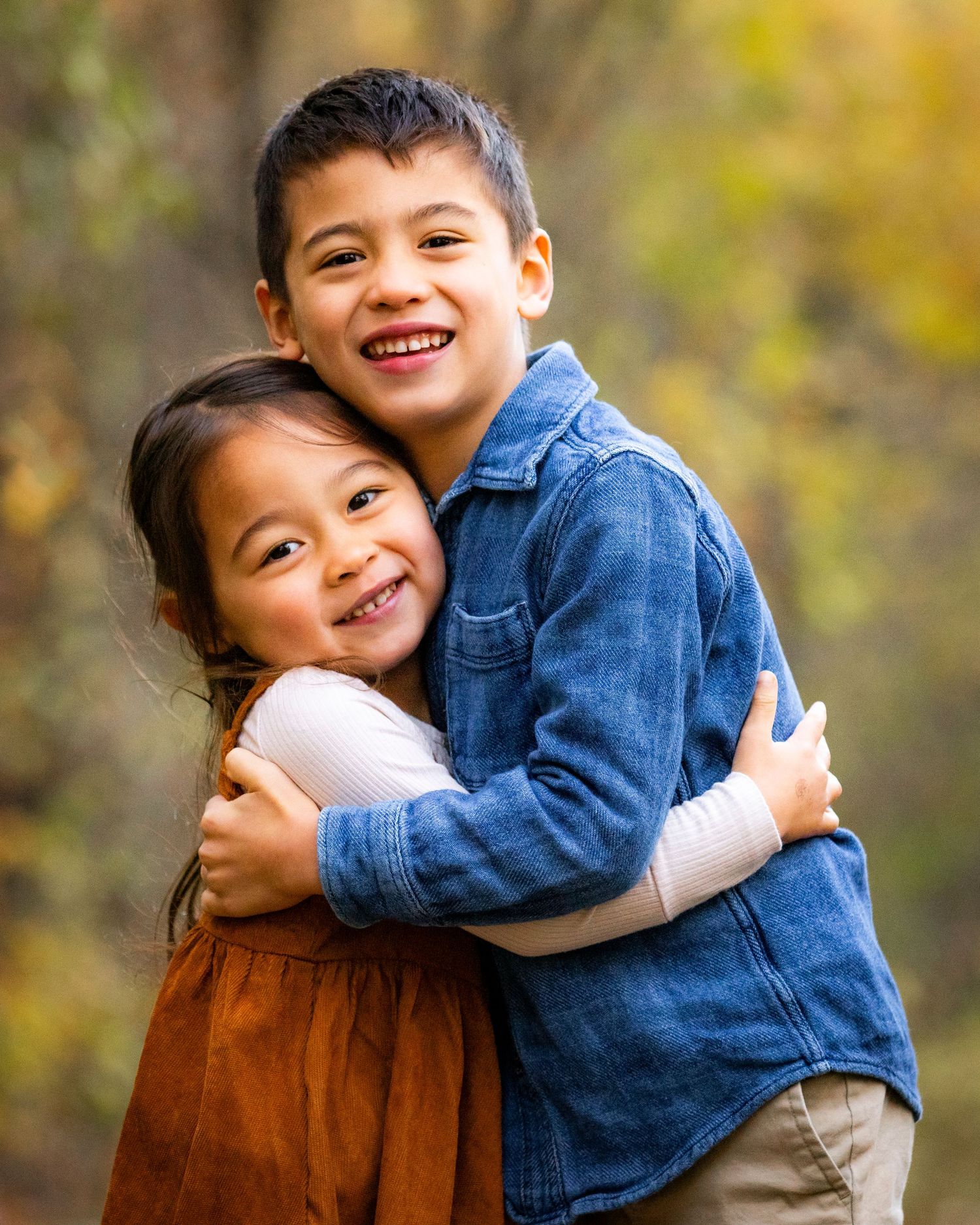 A young brother and sister share a warm hug while smiling outdoors during an autumn family photo session.
