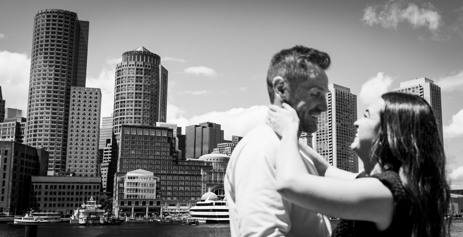 Black and white photo of a couple embracing with Boston skyline and harbor in the background.